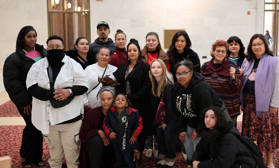 A diverse group of people, including adults and children, stand together indoors on a patterned carpet, facing the camera.