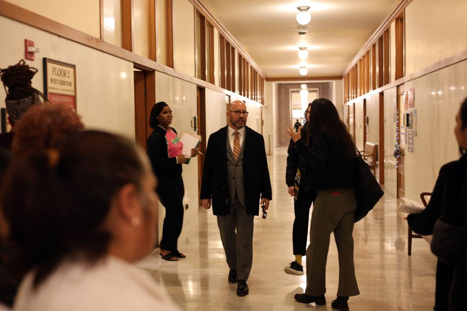 A man in a suit walks down a hallway lined with people, some standing and talking, in what appears to be an office or government building.