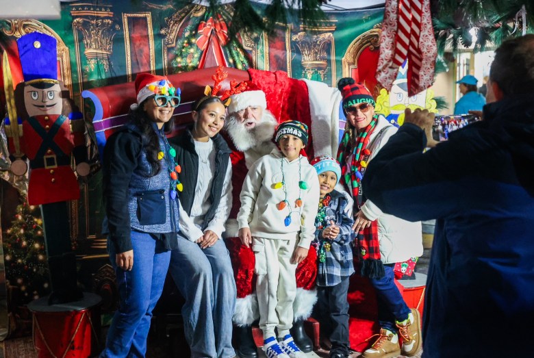Santa on a throne surrounded by five people dressed up in winter gear, wit three of the people wearing holiday lights necklaces.