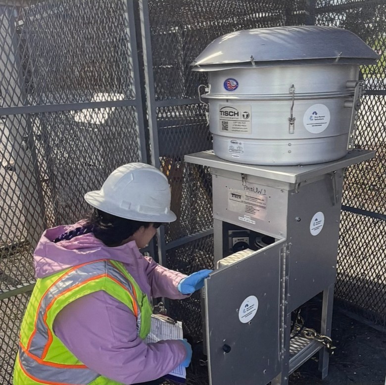 a person inside a metal cage wears safety gear and inspects a metal cabinet topped with a large cylinder