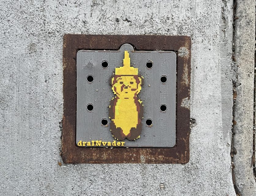 A pixel art sticker of a yellow peanut with a pointed top is attached to a storm drain; the text "drAINvader" appears in yellow at the bottom left.
