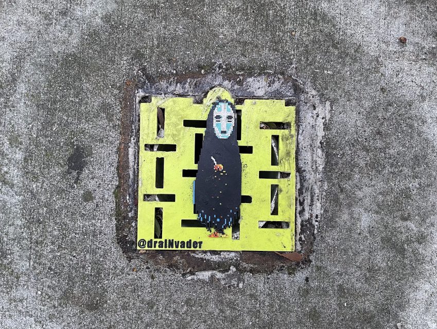 A street drain cover painted bright yellow with a pixel art figure resembling No-Face from Spirited Away; "@dralIVvader" is written on it.