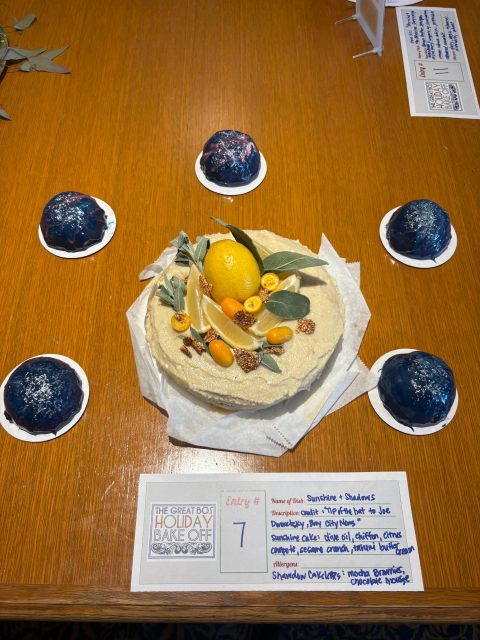 A decorated cake topped with lemon, kumquats, figs, and leaves sits on a table, surrounded by six dome-shaped desserts; an entry card labeled “Entry #7” is placed in front.