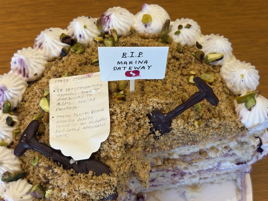 A partially sliced cake decorated with piped cream, pistachios, chocolate hammers, and signs reading “R.I.P. Marina Gateway” and a handwritten note detailing amendments and shortages.