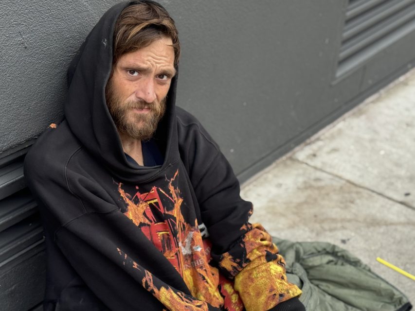 A man with a beard and brown hair sits on the sidewalk against a gray wall, wearing a black hoodie with an orange design and looking at the camera.