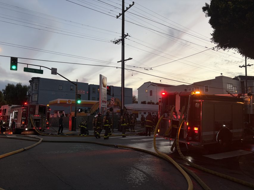 Fire trucks and firefighters respond to an emergency at dusk on a city street, with hoses stretched across the road and lights flashing.