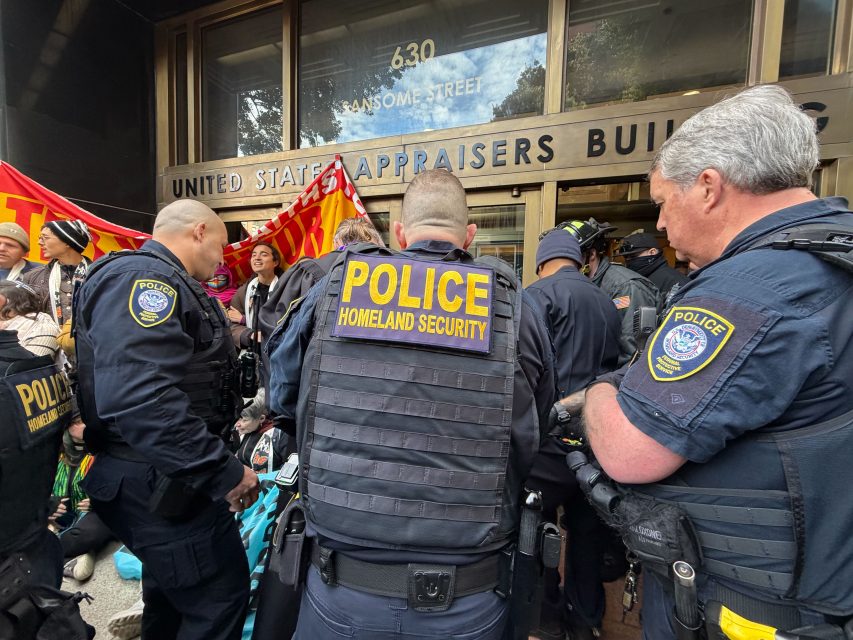 A group of Homeland Security police officers stands in front of the "UNITED STATES APPRAISERS BUILDING" amid a crowd and red banners during an immigration-related gathering.