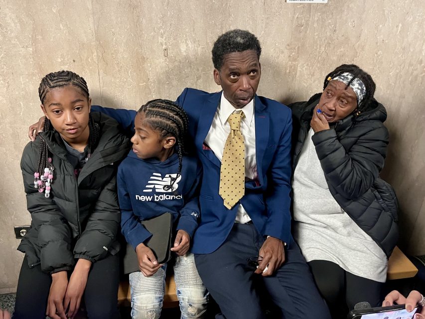 Four people sit close together on a bench indoors; an adult man in a suit is flanked by two girls and an older woman who is wiping her eye.