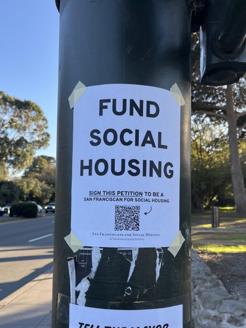 A sign taped to a pole reads "Fund Social Housing" and encourages people to sign a petition, with a QR code and a park visible in the background.