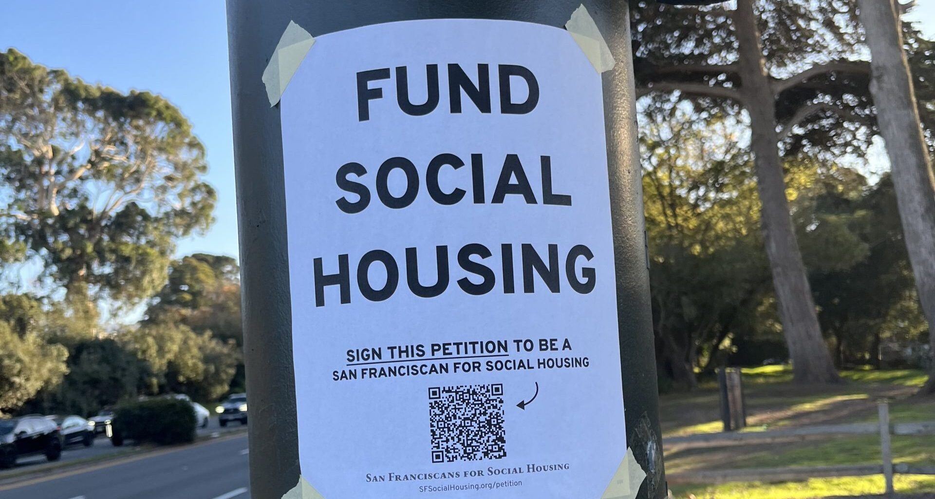 A flyer taped to a pole reads "Fund Social Housing" and urges people to sign a petition, with another notice below about public transit funding. Trees and a roadway are in the background.