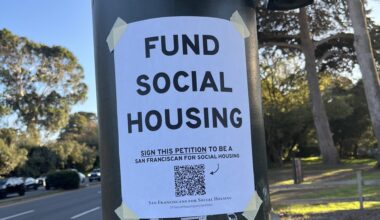 A flyer taped to a pole reads "Fund Social Housing" and urges people to sign a petition, with another notice below about public transit funding. Trees and a roadway are in the background.