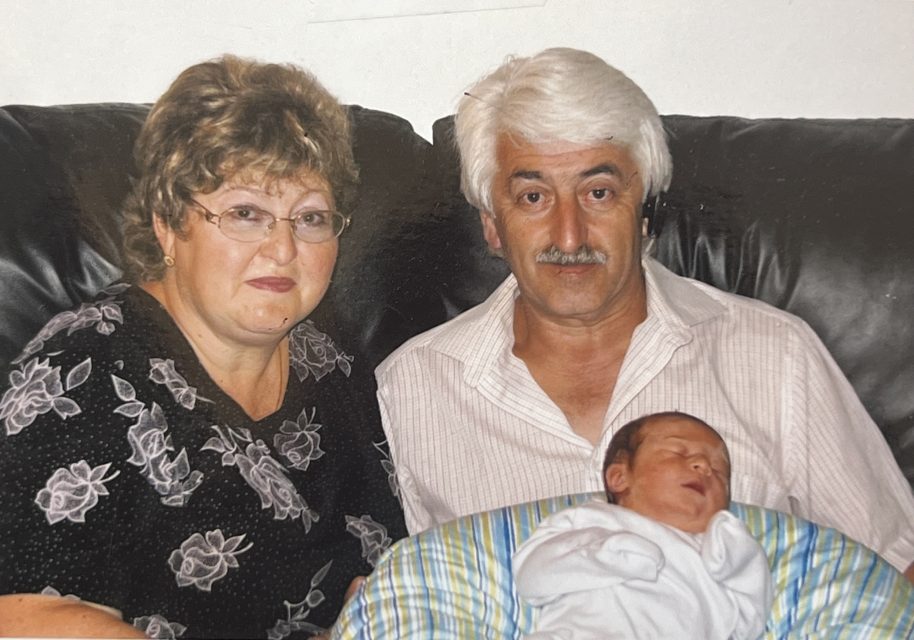 An older woman and man sit on a dark couch, the man holding a sleeping newborn wrapped in a blanket on his lap.