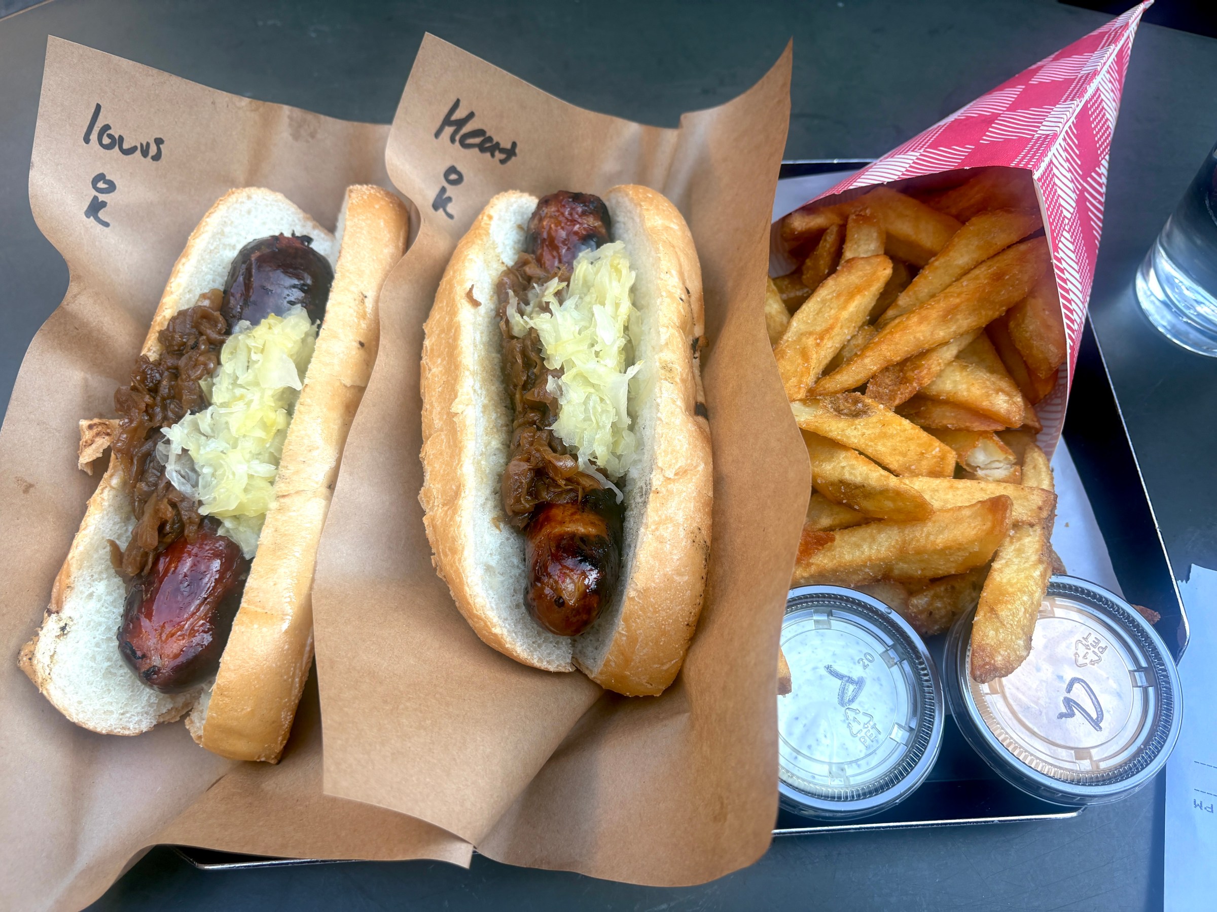 Tray with two sausages topped with sauerkraut on soft rolls with a side of Belgium fries and dipping sauce.