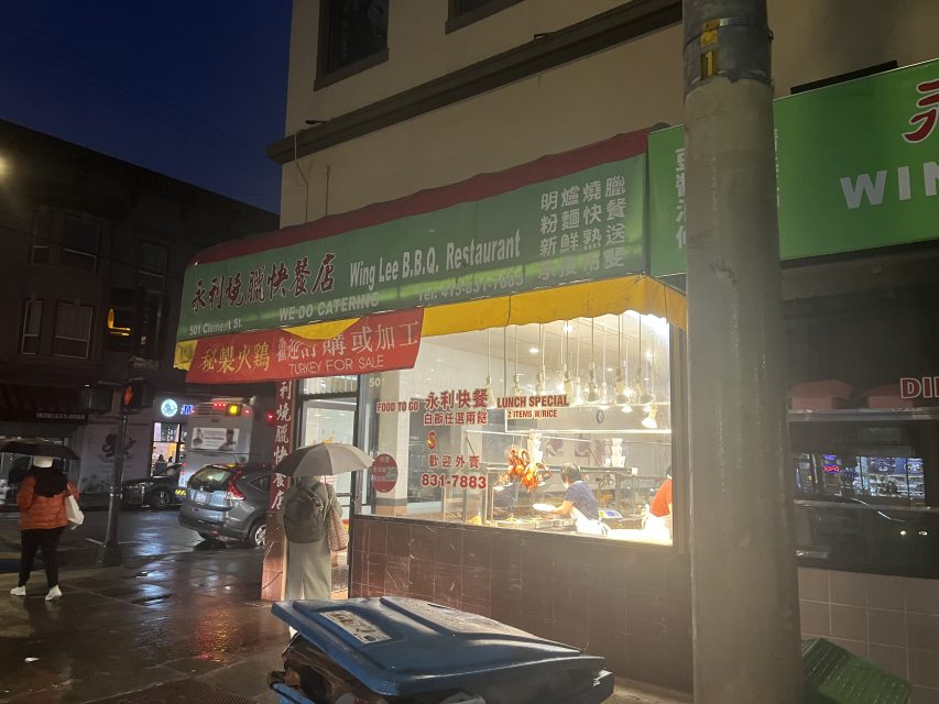 Wing Lee B.B.Q. Restaurant storefront at night, with lights on inside, "For Sale" and "Lunch Special" signs visible, people walking by, one holding an umbrella.