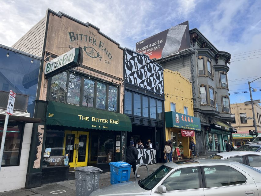 Street view of The Bitter End bar and grill, neighboring buildings with murals, parked cars, and people walking on a cloudy day.