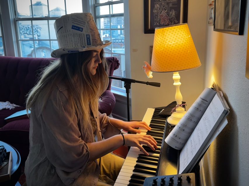 A person wearing a newspaper-patterned hat plays an electric keyboard next to a lit table lamp with sheet music open in a cozy room, glancing at parking tickets scattered on the nearby desk.