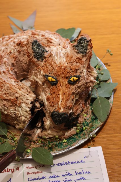 A round cake decorated to resemble a fox's head, with detailed icing for fur and eyes, placed on a plate with green leaves around it. A knife is partially cutting the cake.