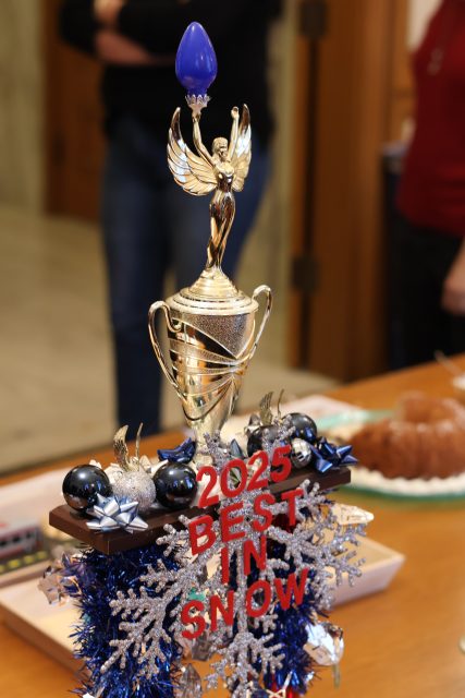 A gold trophy with a winged figure, blue decorations, and “2025 BEST IN SNOW” text stands on a table near a bundt cake and other items.
