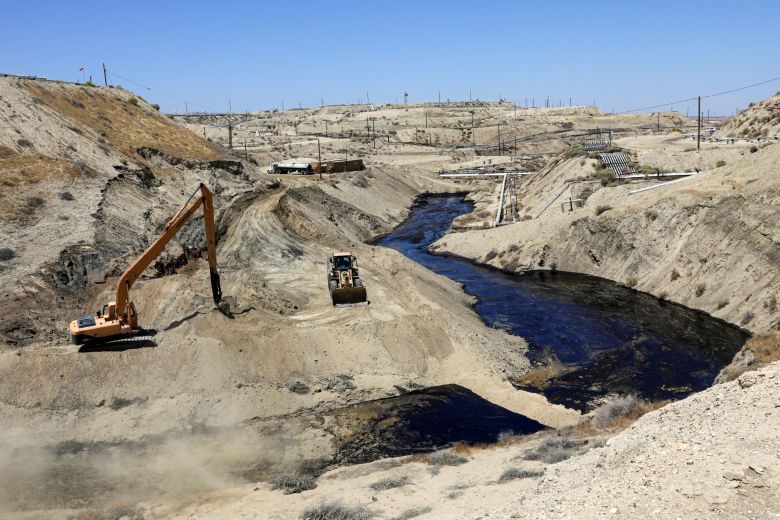 Construction vehicles work near an oil spill in a barren, hilly landscape under a clear blue sky.