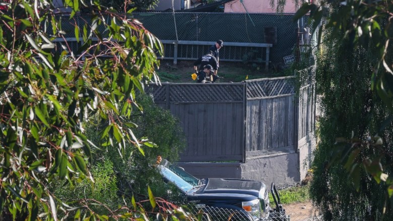 Two people in NCIS uniforms near a wooden fence with a black pickup truck in front, surrounded by trees.