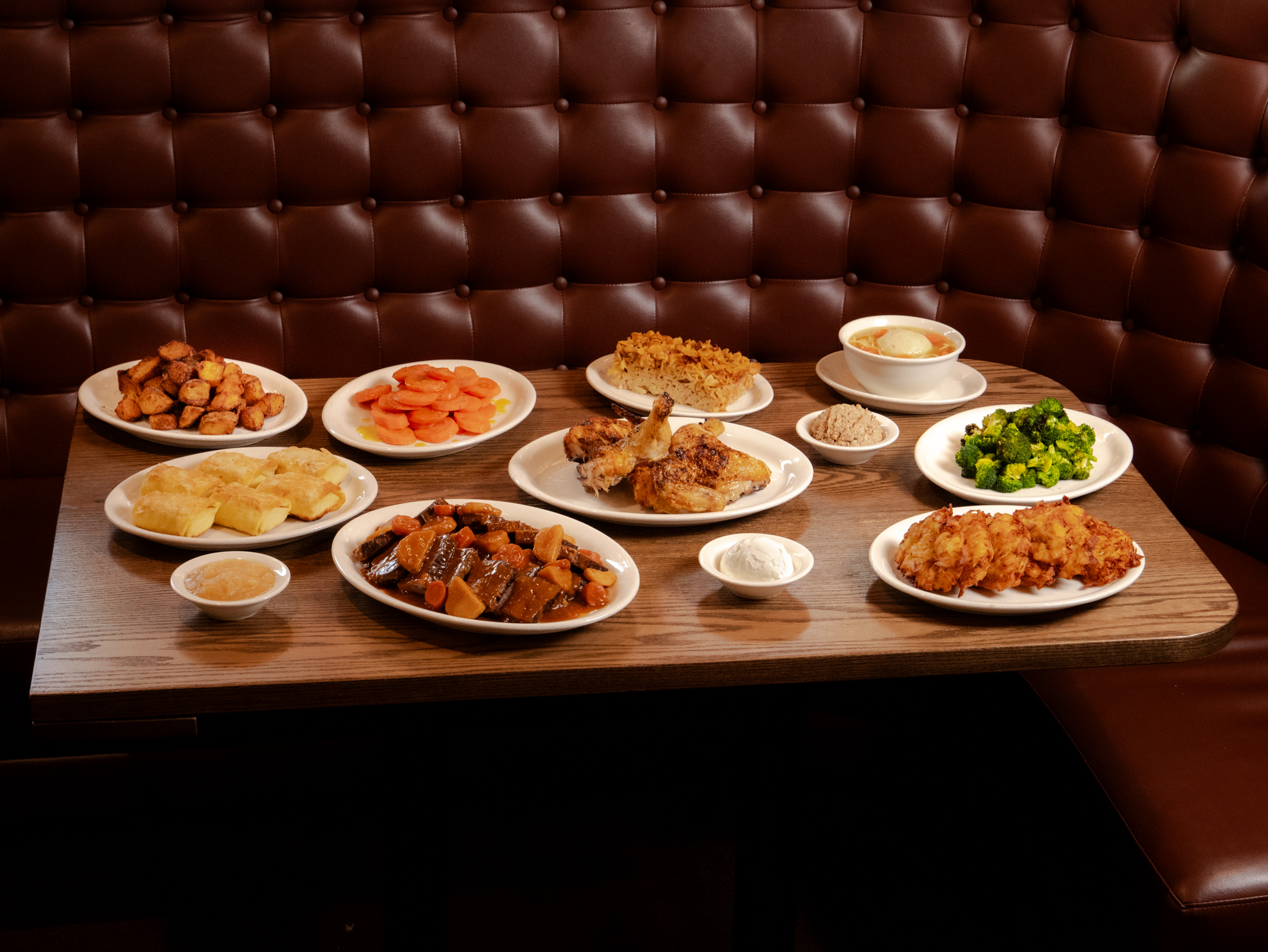 Large wood table covered with traditional Hanukkah celebration dishes (including latkes), all against a plush leather banquette backdrop.