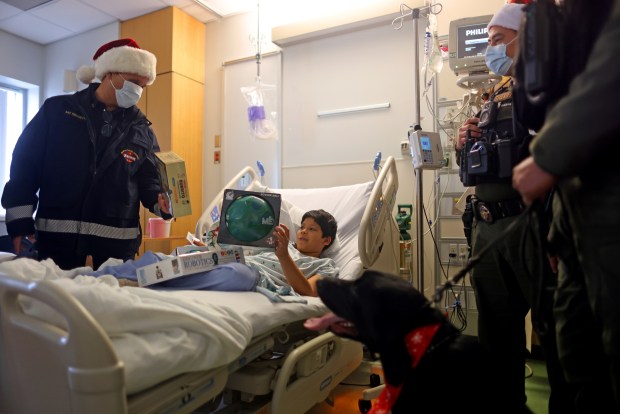 Art Tomasetti, left, with the Santa Clara County Fire Department delivers Christmas gifts along with members of the Santa Clara County Sheriff's Department to patient Kevin Fernandez in the children's wing of the Santa Clara Valley Medical Center on Wednesday, Dec. 17, 2025, in San Jose, Calif. The two departments collected hundreds of toys from the community to be distributed at the hospital and other sites throughout the county. (Aric Crabb/Bay Area News Group)
