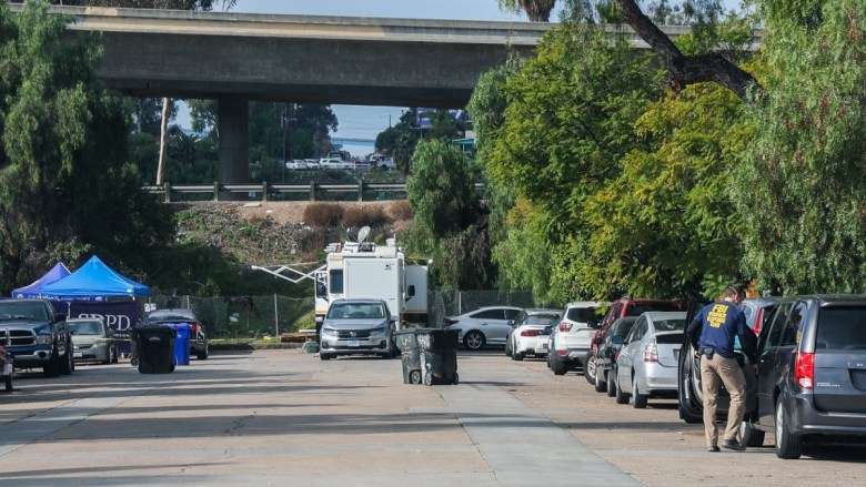 A street under an overpass with parked cars, a police canopy tent, a mobile command vehicle, and a person in an FBI jacket.