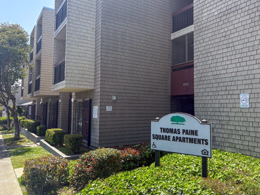Exterior view of Thomas Paine Square Apartments building with a sign in front, shrubbery, and accessible entrance symbols visible.