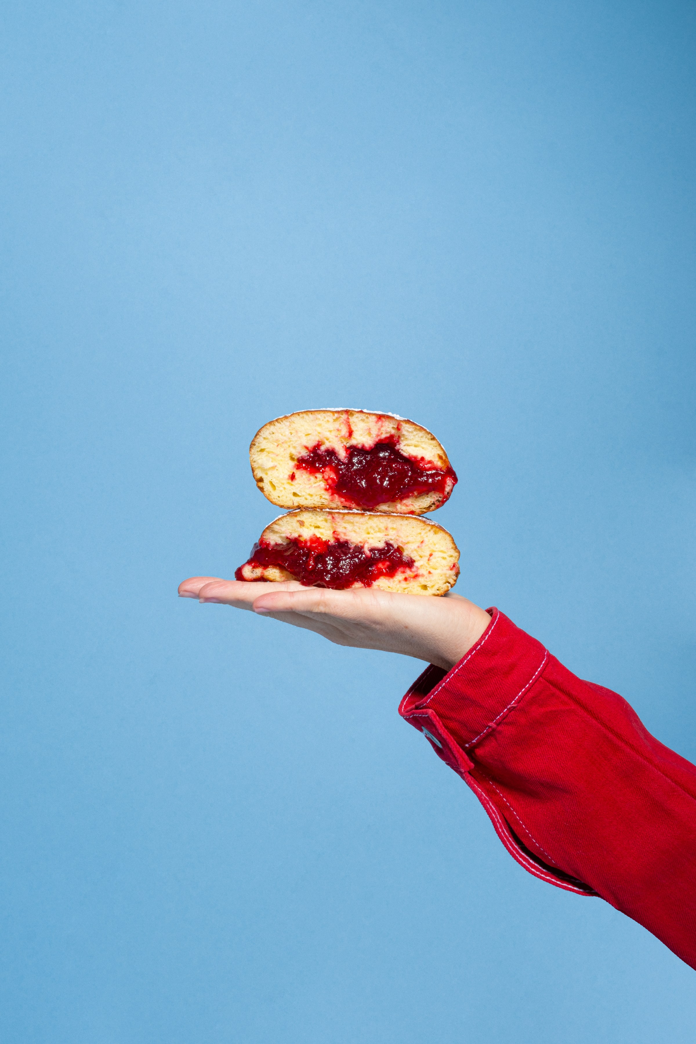 An outstretched hand and arm in a red jacket holding up jam-filled sufganiyot against a bright blue backdrop.