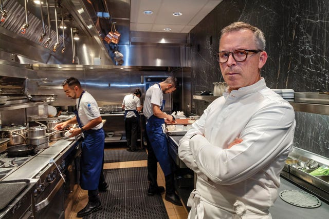 chef william bradley stands arms crossed inside the addison kitchen, while his culinary team plates a multi course dinner in the background.