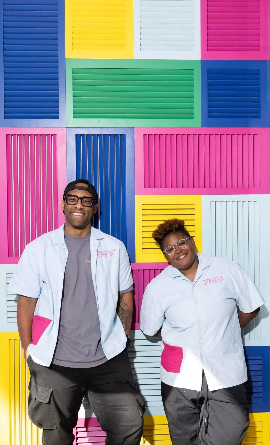 Two people pose for a portrait against colorful blinds.