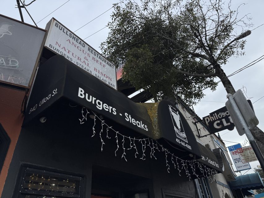Street view of a restaurant with signs reading "Burgers - Steaks" and "Bull's Head Restaurant," strings of lights on the awning, a tree overhead, and a warm sunset glow illuminating the scene.