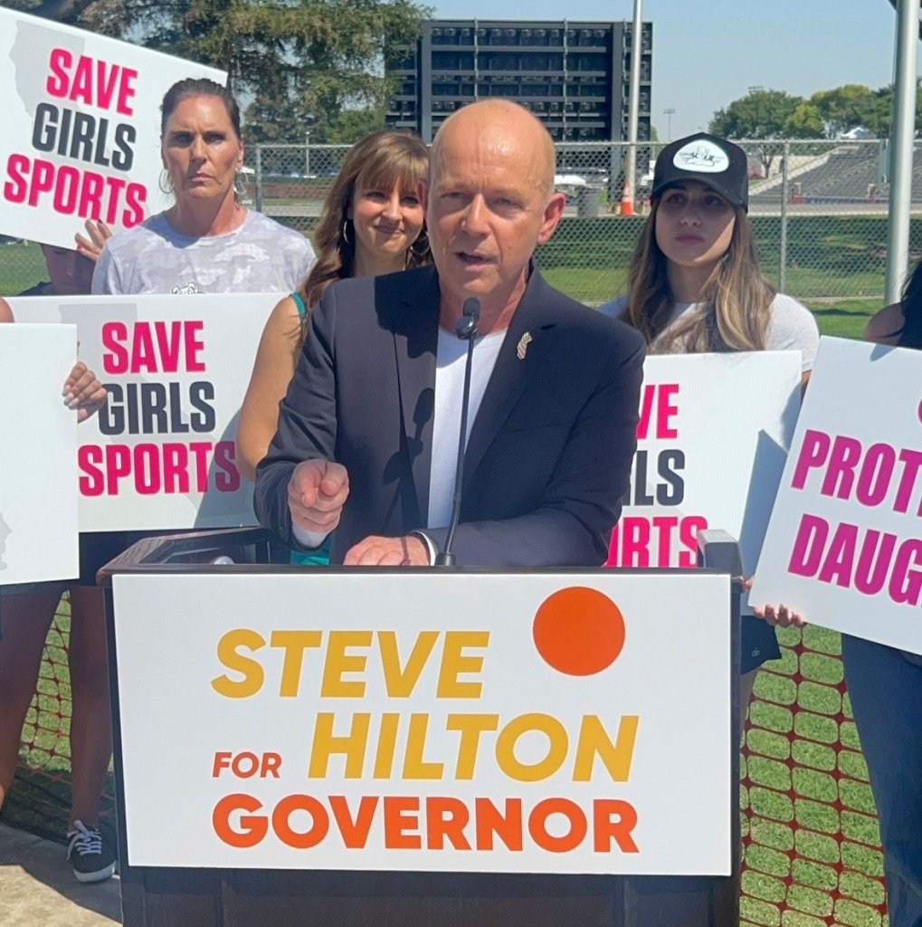 California gubernatorial candidate Steve Hilton speaks at a podium with "Steve Hilton for Governor" written on it, while people behind him hold signs reading "SAVE GIRLS SPORTS."