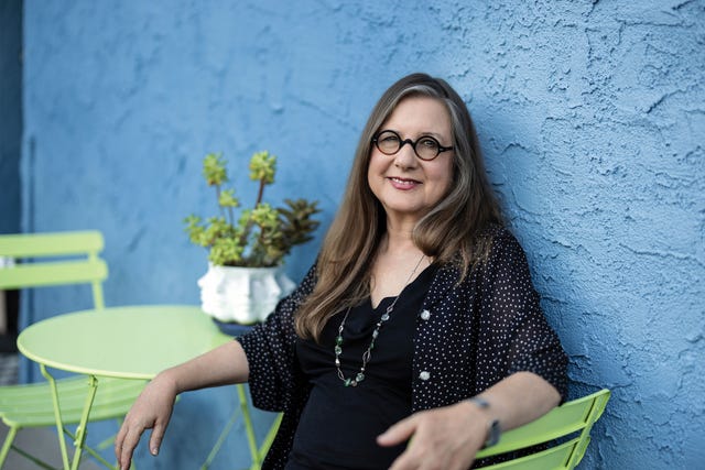 janet fitch seated outdoors against a blue wall, smiling warmly, with a vase of yellow flowers behind her—capturing her reflective, creative spirit.