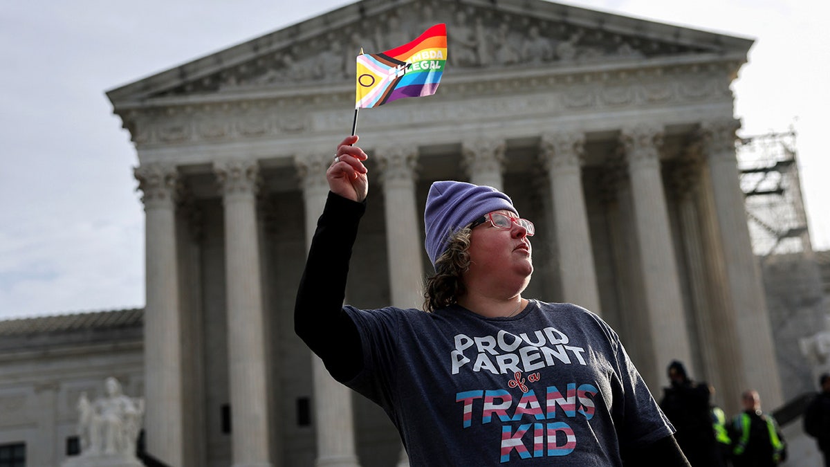Parent of transgender child holds up the LGBTQ+ flag in front of the Supreme Court building.