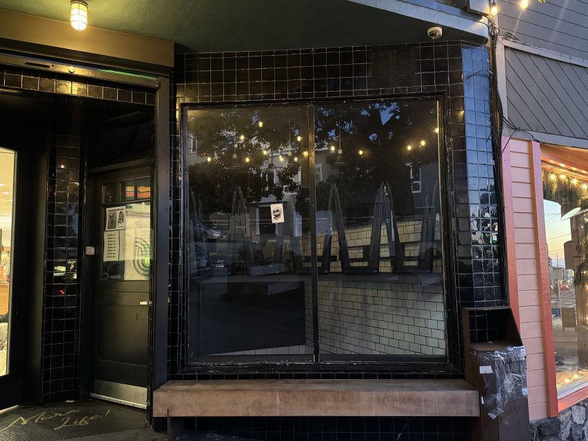 A dark, empty storefront with black tile exterior, stacked chairs inside, and two notices posted on the window, photographed at dusk.