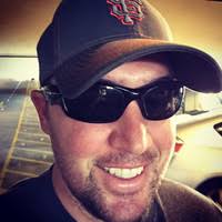 A man wearing a San Francisco Giants baseball cap and dark sunglasses smiles while standing in a parking garage.