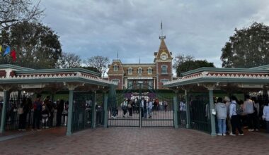 A large crowd of guests in line outside the gates waiting to enter Disneyland Park in Southern California with the Main Street station of the Disneyland Railroad in the background with cast members.