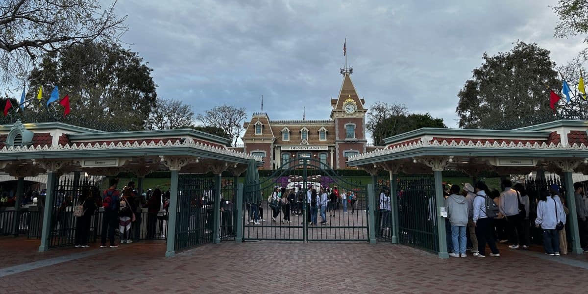 A large crowd of guests in line outside the gates waiting to enter Disneyland Park in Southern California with the Main Street station of the Disneyland Railroad in the background with cast members.
