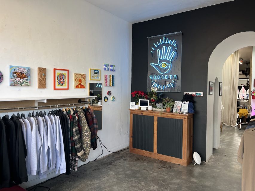 Clothing racks and art display on a white wall, counter with plants and a neon "Society" sign, and an arched entryway in a modern boutique.