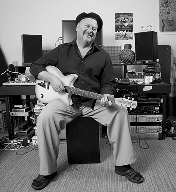 A man sitting on a stool in a home studio, playing an electric guitar and smiling, wearing a hat, loose pants, and sandals.
