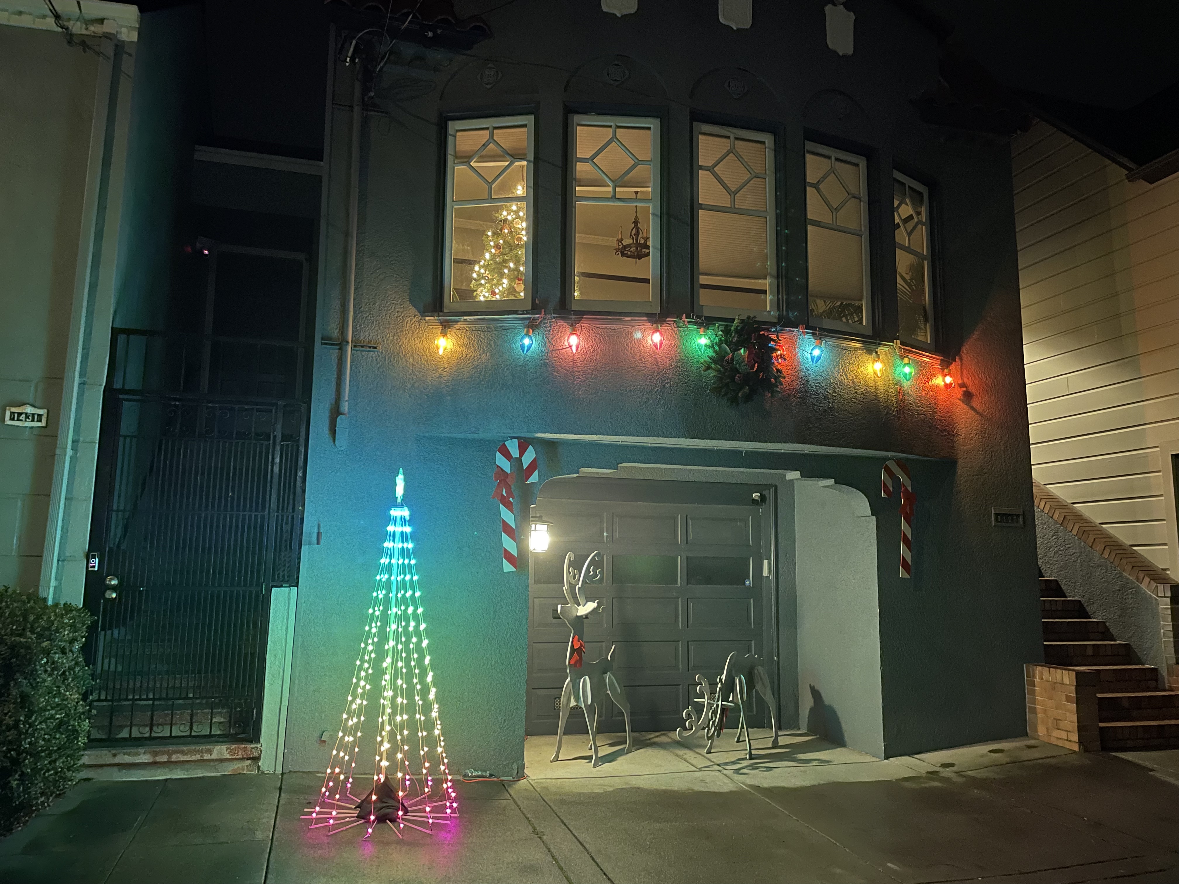 A house decorated at night with multicolored lights, a lit outline of a Christmas tree, two reindeer figures, and candy cane decorations by the garage.