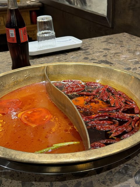 A divided hot pot with spicy broth containing red chili peppers on one side and a milder red broth on the other, next to a glass bottle of Coca-Cola on a marble table.