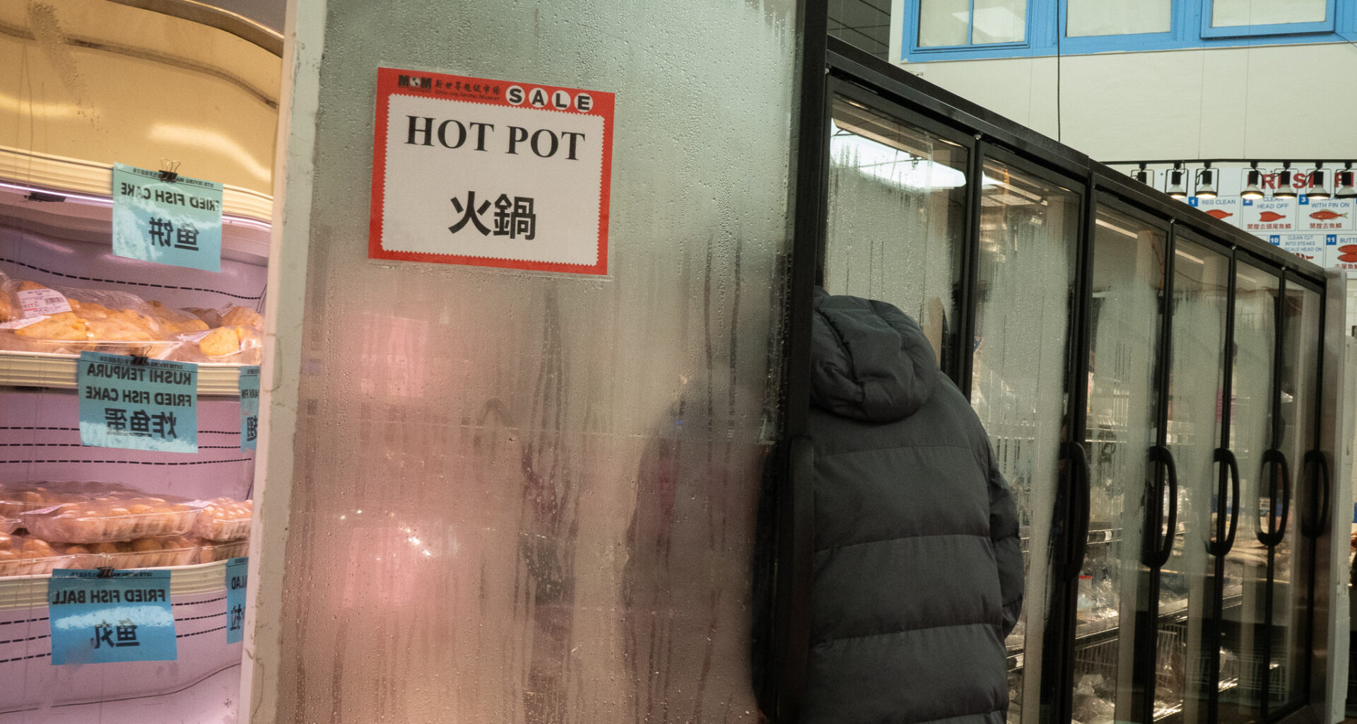 A person wearing a dark coat stands partly inside a foggy glass freezer door labeled "HOT POT" in a grocery store. Shelves of packaged food are visible to the left.