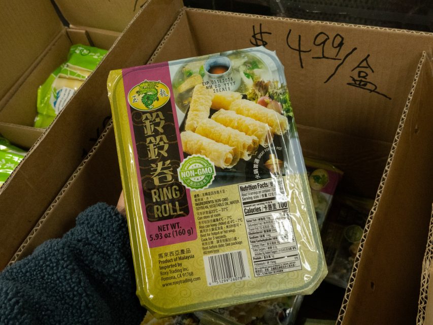 A hand holds a package of non-GMO ring rolls in front of a box labeled $4.99 per box. The package label displays nutrition facts and product details.