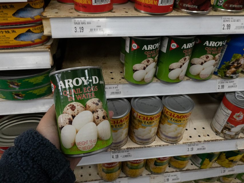 A hand holds a can of Aroy-D quail eggs in water in front of similar cans on a grocery store shelf.