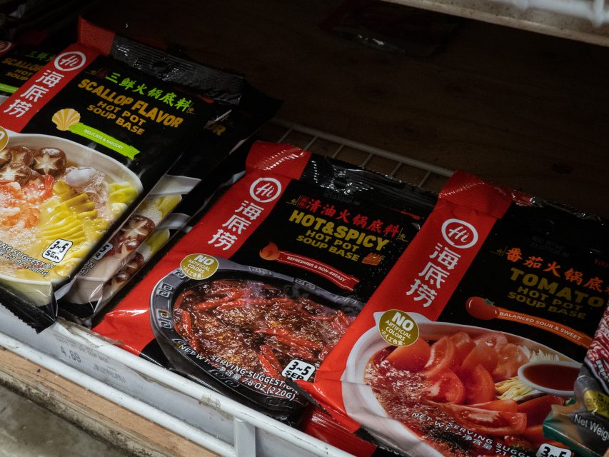 Three different hot pot soup base packets—scallop flavor, hot & spicy, and tomato flavor—are displayed on a store shelf.