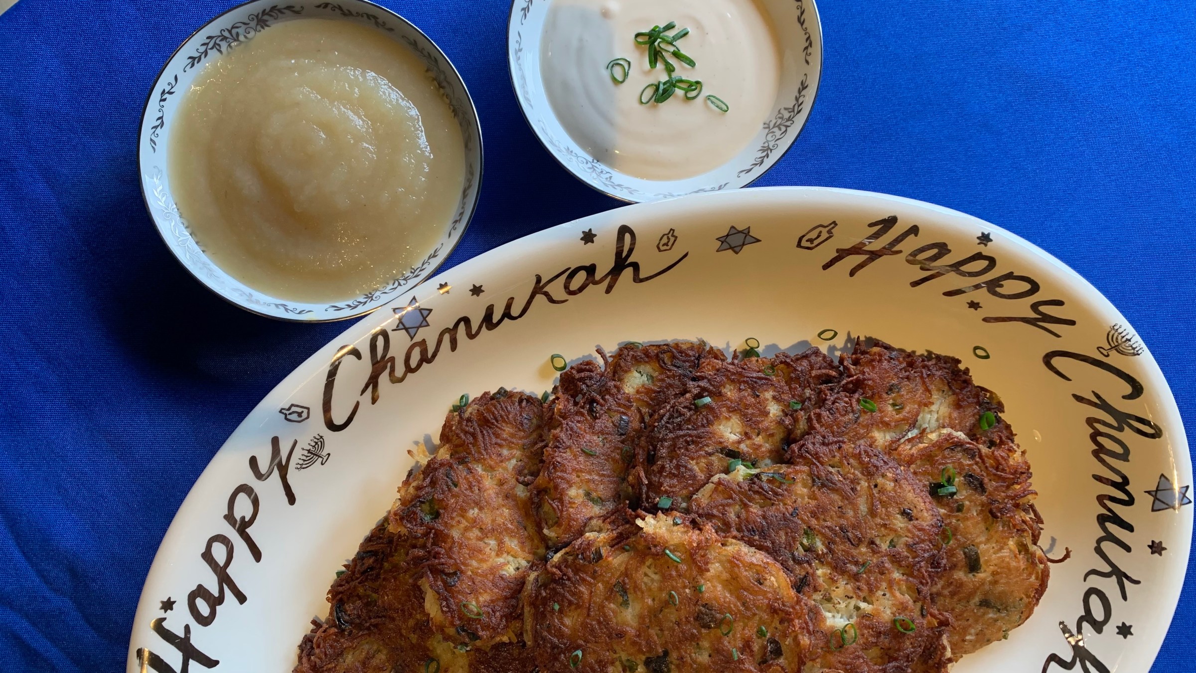 Bird-eye-view of a decorative plate of deeply browned latkes flanked by ramekins of applesauce and vegan (cashew) sour cream.