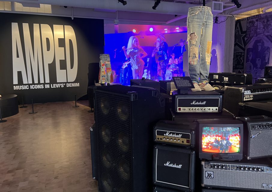 Room with a large "AMPED: Music Icons in Levi's Denim" mural, band performing on stage, and stacked Marshall amplifiers with jeans and a small TV in the foreground.