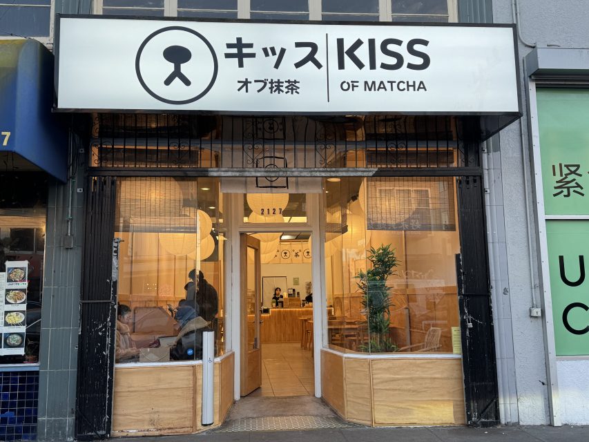Street view of a cafe named "Kiss of Matcha" with large front windows, minimal wooden decor, and a prominent sign in English and Japanese above the entrance.